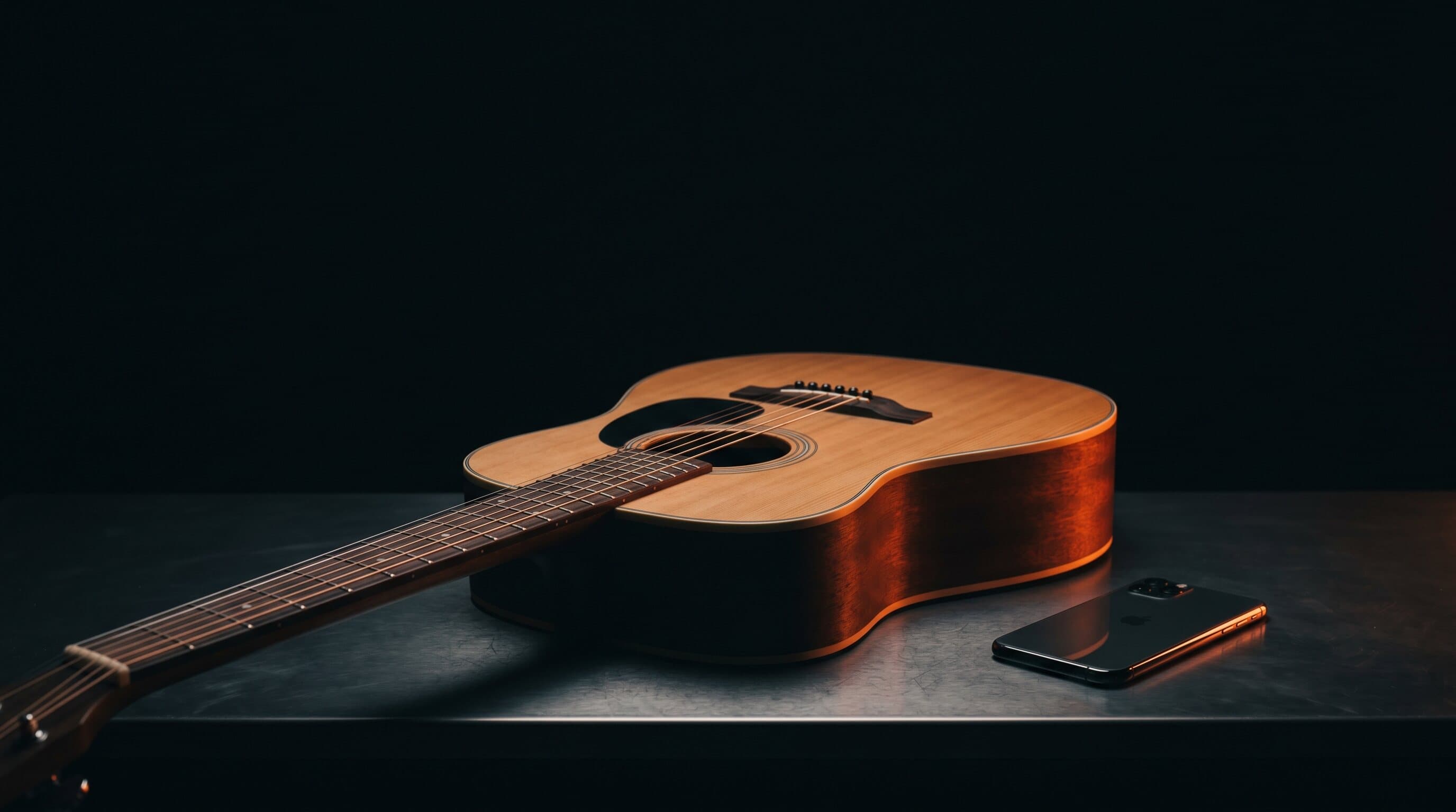 A steel-string acoustic guitar and a smartphone on a dark studio surface with warm orange lighting