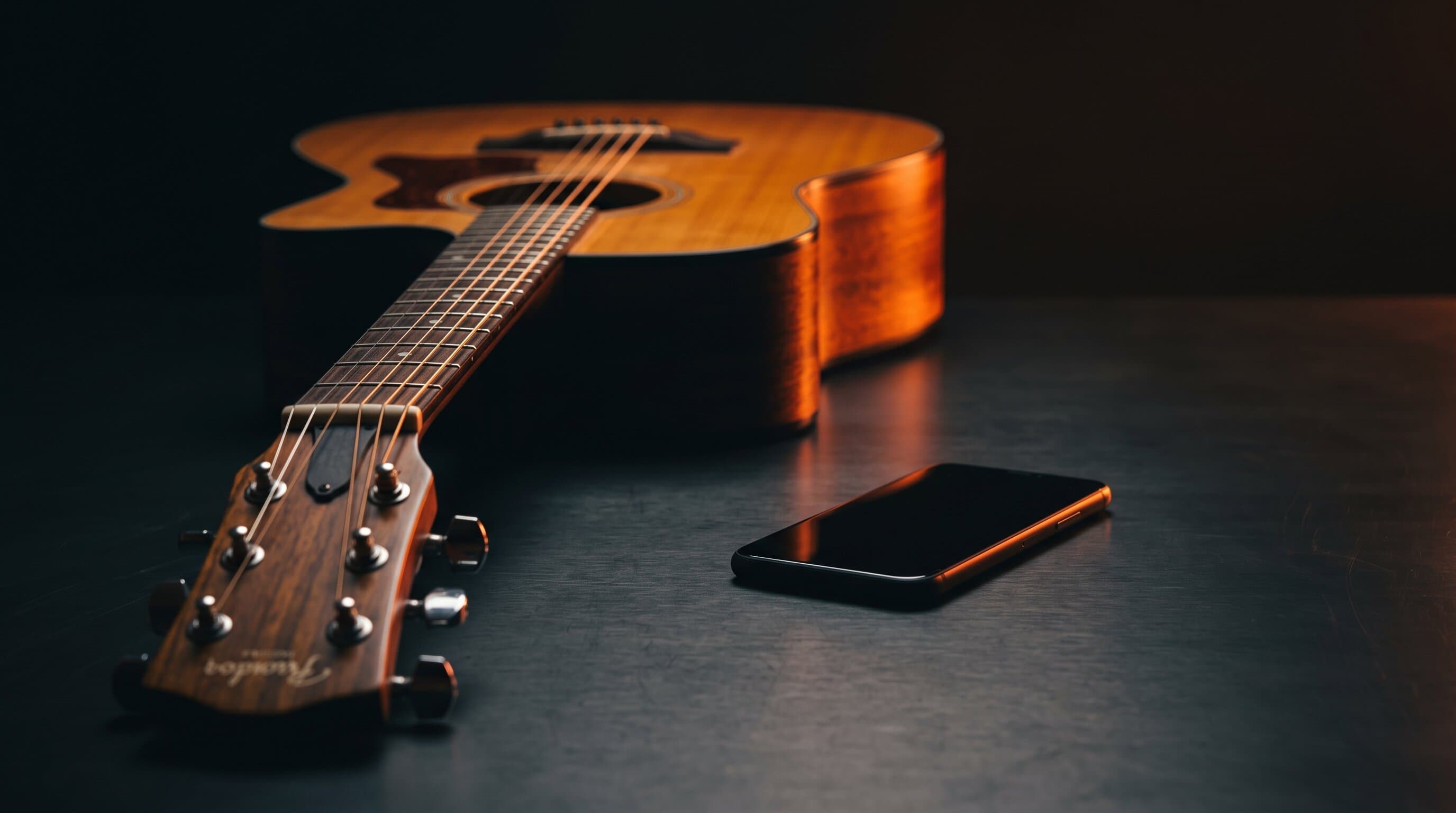 An acoustic guitar resting on a dark studio desk beside a smartphone, lit with warm orange light, representing the JustinGuitar practice app