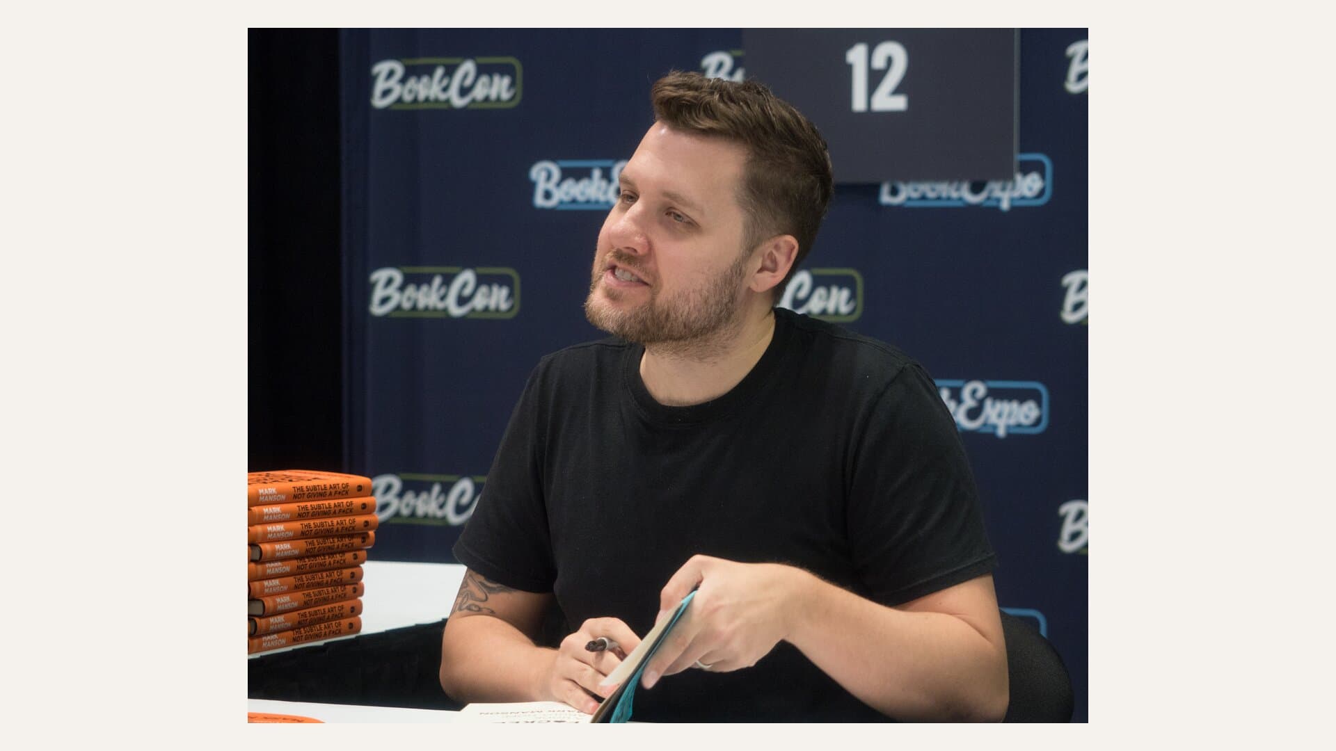 Mark Manson signing books at BookCon in 2019, beside a stack of orange copies of The Subtle Art of Not Giving a F*ck