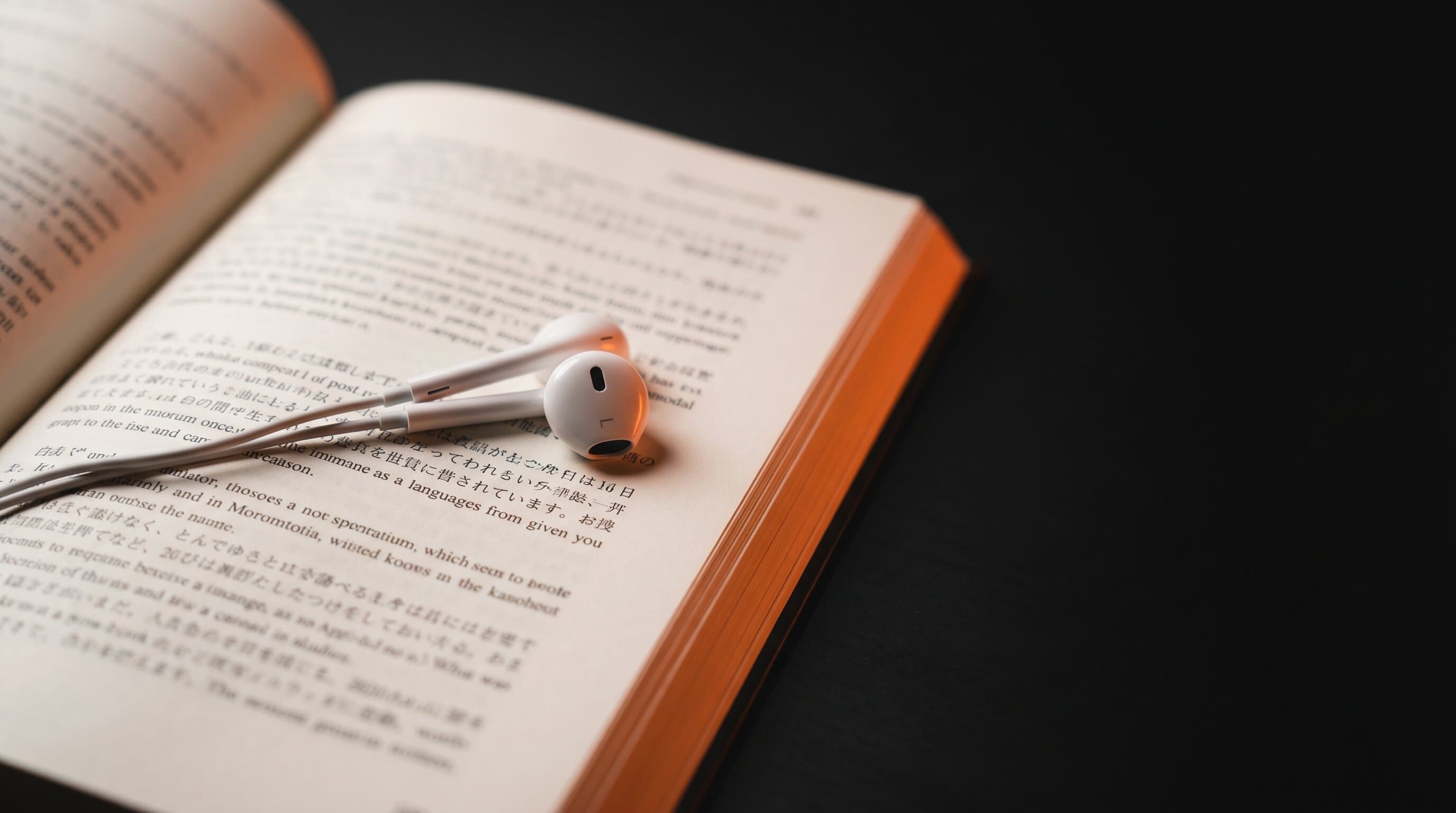 White earbuds resting on an open book of foreign-language text, lit with a warm orange accent on a dark surface